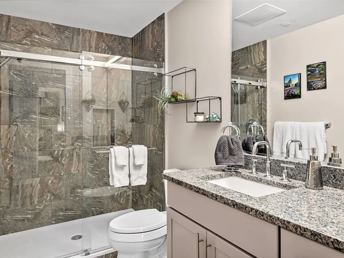 A sophisticated bathroom with a glass shower enclosure, granite countertop, and decorative shelving with plants, suffused with morning light—suggesting a space designed for refreshing routines and luxurious relaxation.
