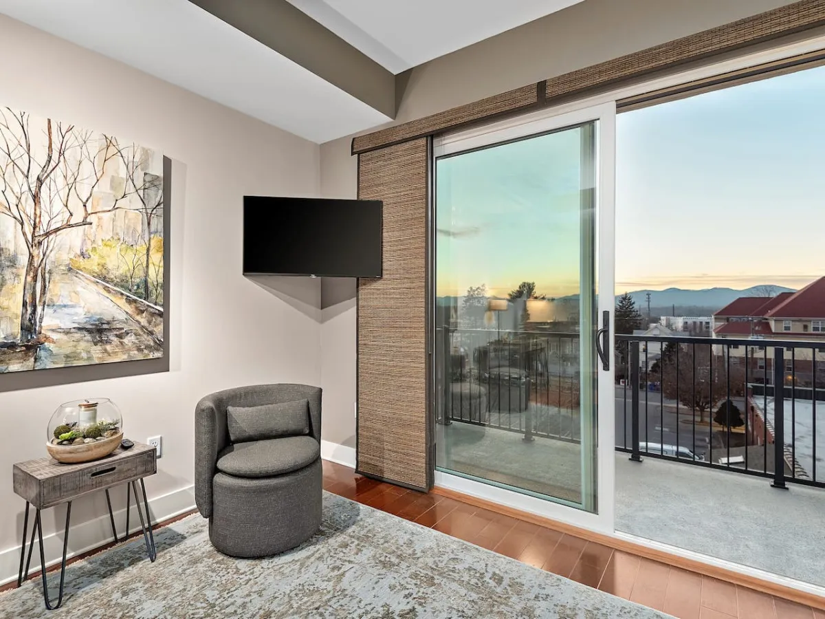 This image captures a luminous living room with plush seating, striking artwork on the wall, and a cozy chair inviting relaxation. The room extends to a stylish balcony that offers sunset views of the city, blending indoor comfort with outdoor allure. The scene feels calm and serene, with natural light pouring in, highlighting the open connection to the balcony and the cityscape beyond.