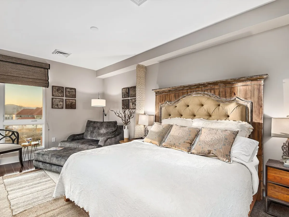 Another king bedroom with a tufted headboard, plush armchair, soothing color palette, and decorative wall art, offering tranquil views through large windows and a cozy reading nook to unwind in after a long day.