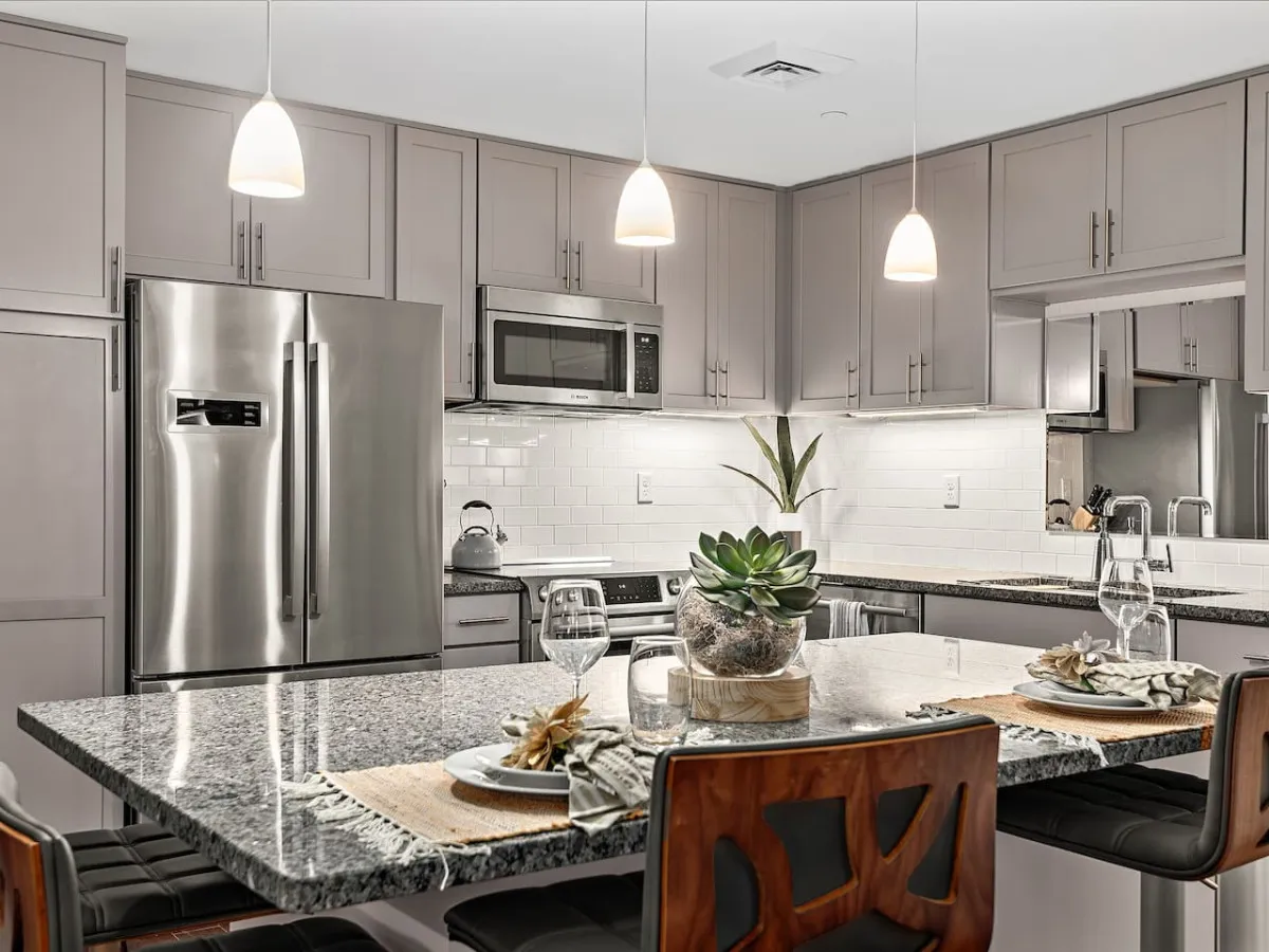 A stylish kitchen featuring a granite countertop, modern high-end appliances, and elegant hanging light fixtures. A succulent centerpiece sits atop the island, creating a warm and inviting gathering space. The room exudes a welcoming atmosphere, ideal for cooking, entertaining, and enjoying meals together.