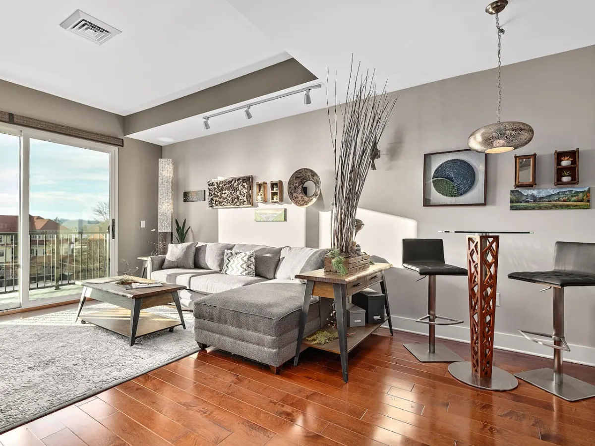 A stylish open-concept space with a comfortable sofa, a sleek bar table, decorative wall art, and expansive windows that flood the area with light. The contemporary setting feels both lively and secure, reflecting the blend of city energy and peaceful retreat mentioned in the section.
