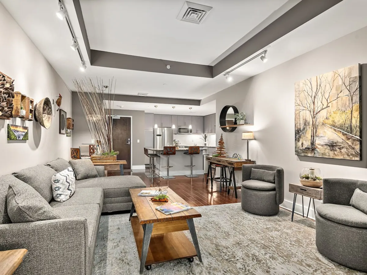 The image showcases a bright, contemporary living area featuring a plush sofa, stylish coffee table, vibrant local artwork, and a sleek bar area. Sunlight fills the inviting space, creating a perfect setting for relaxing, entertaining, or sharing memorable moments with friends and family.