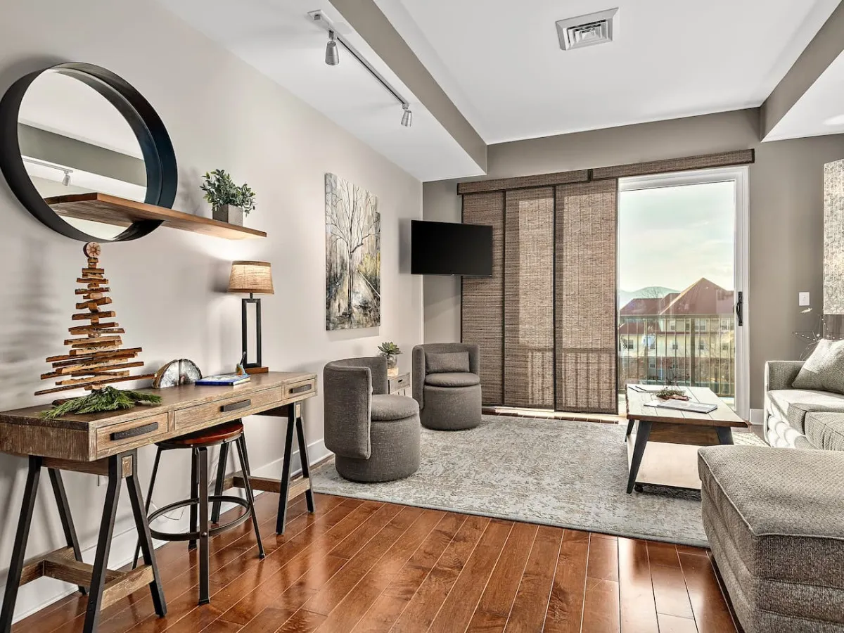 A welcoming entryway area featuring a wooden desk with a small decorative tree, a circular mirror above, tasteful wall artwork, and a cozy seating area with a sofa. A sliding glass door offers a glimpse of the outside, enhancing the sense of arrival.