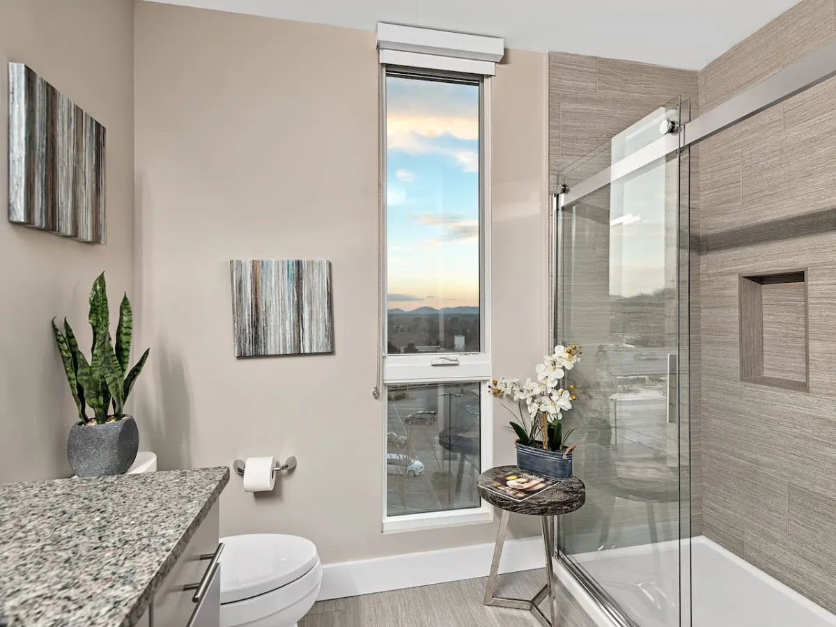 This image features a large window offering a scenic view, with glimpses of the cityscape and surrounding mountains visible in the distance. The space is accentuated by framed wall art and a decorative plant, creating a tranquil nook ideally suited for unwinding after a day out. The modern vanity with granite countertop and a glass shower enclosure are also visible, giving an elegant touch to the scene.