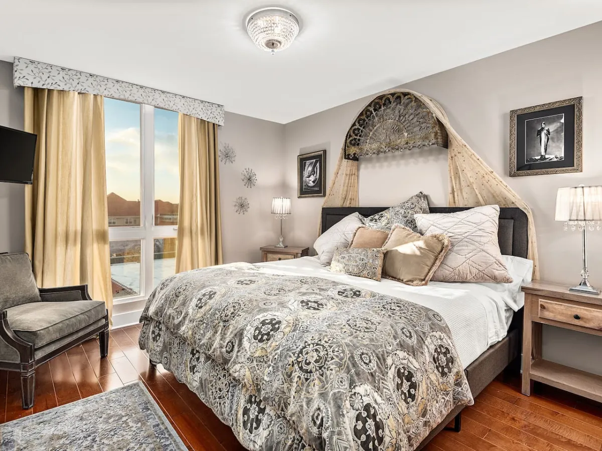 A king bedroom featuring a plush bed with decorative bedding, a large window draped with curtains that bathes the room in soft natural light, tasteful artwork on the walls, and a cozy armchair in the corner—evoking a sense of inviting comfort.