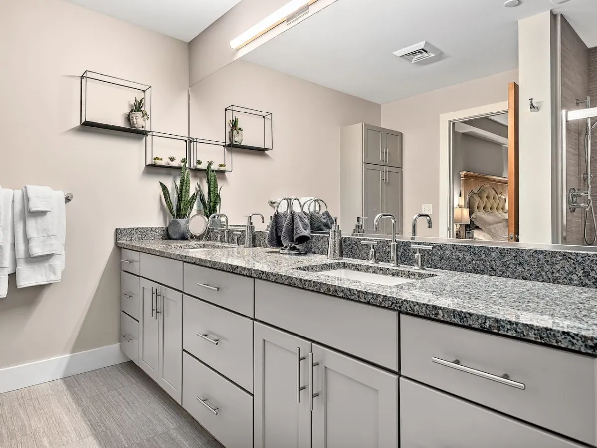 A modern bathroom scene focused on a granite countertop, stylish faucet fixtures, and decorative shelving with lush plants. Towels hang on a rack, and a large mirror reflects more of the organized, serene space. Soft lighting and subtle décor convey a calming, spa-like environment perfect for relaxation after a day out.